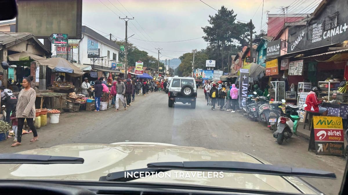 Market stalls and shopfronts lining a busy Malagasy town street, seen from behind a car windscreen