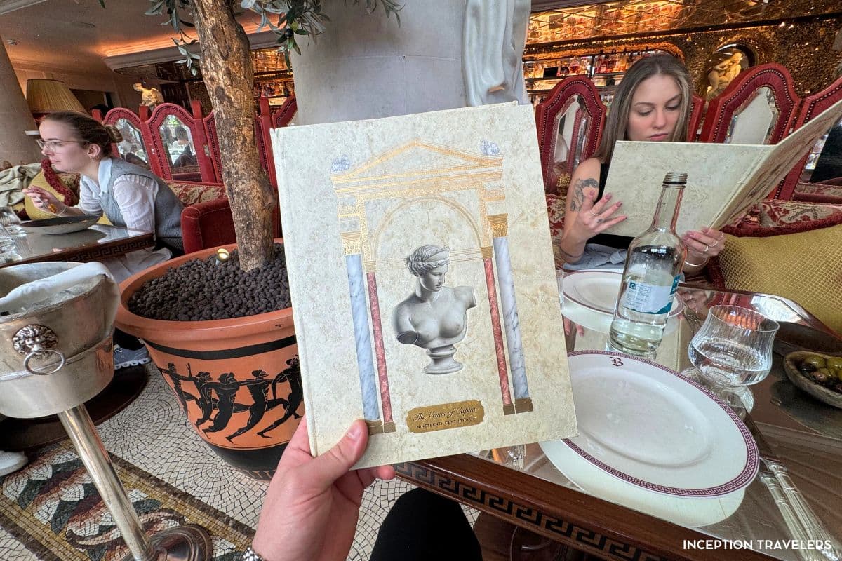 Marble-effect Venus of Capua menu held up at a Bacchanalia table, with the gold mosaic bar behind