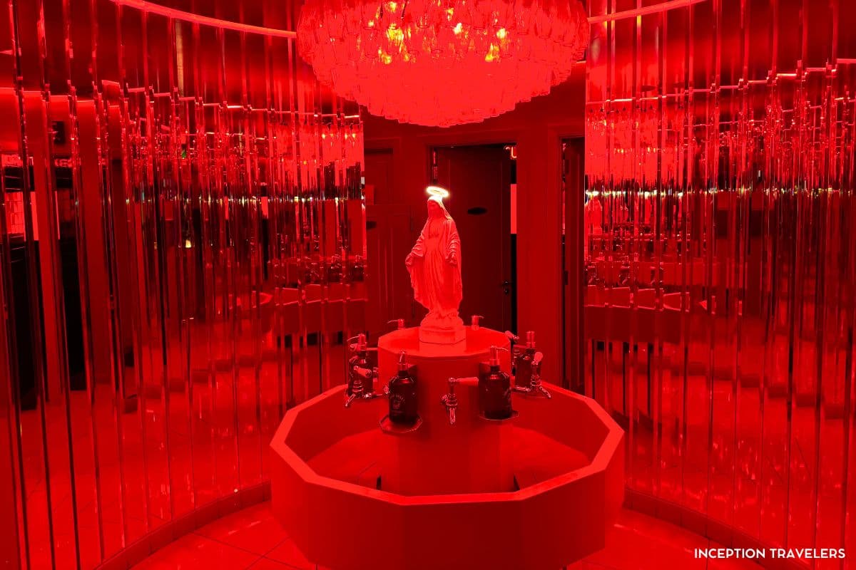 Red-lit circular washroom with mirrored walls, a chandelier and a haloed Madonna statue above a ring of soap dispensers