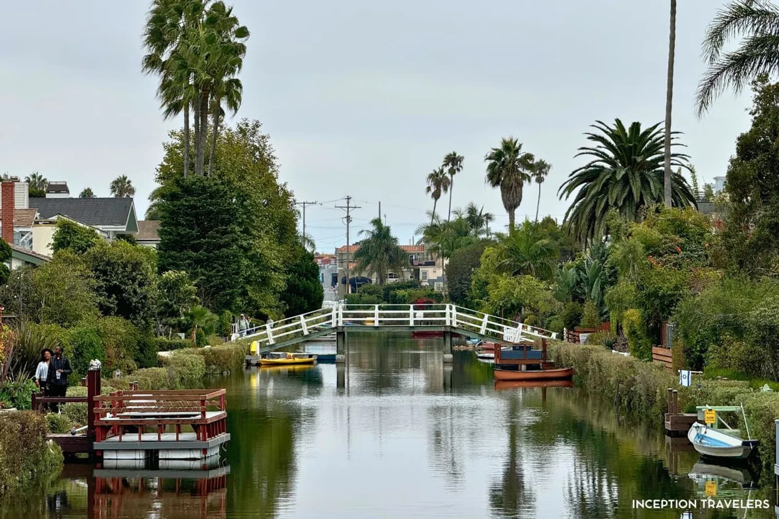 White footbridge arching over a calm Venice Canal lined with moored boats, gardens and palms